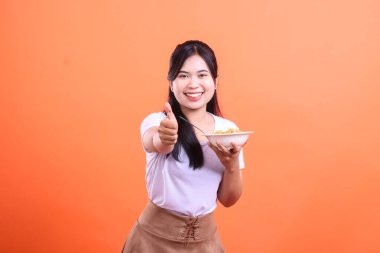 Bir kase lezzetli erişteyle gülümseyen genç bir kadın, en iyi yemek deneyimini göstermek için işaret ediyor, turuncu arka planda izole edilmiş.