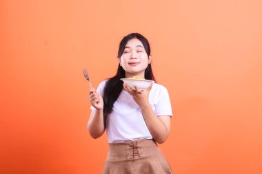 Siyah saçlı genç bir kadın elinde bir kase erişte ve çatal tutarken gözlerini kapatıyor. Bu da yemeğinin tadını çıkardığını gösteriyor. turuncu arkaplanda izole