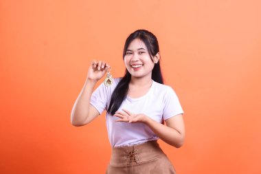 Gülümseyen genç bir kadın, ev sahipliğini ve emlak fırsatlarını temsil eden ev anahtarlarını turuncu arka planda saklıdır..