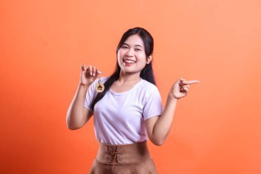 Neşeli genç bir kadın anahtarlık tutuyor ve diğer eliyle yan tarafı işaret ediyor. Gülümsüyor ve turuncu arka planda izole edilmiş yeni evi gösteriyor.