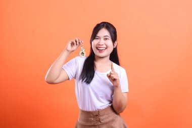 Gülümseyen bir kadın evin anahtarlığını tutuyor, parmağını kaldırıyor, mutlu görünüyor, turuncu arka planda izole edilmiş yeni bir ev ya da mülkün işareti.