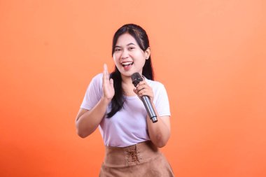 Neşeli genç bir kadın coşkuyla şarkı söylüyor ve mikrofonla el sallıyor, şarkı söylemeye hazır. turuncu arkaplanda izole