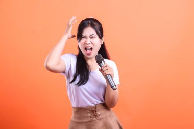A young woman passionately singing or shouting into a microphone with a hand raised in the air, showing a strong expression, isolated on orange background