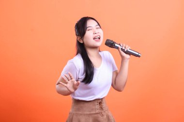 Young woman singing passionately into a microphone, enjoying her hobby, with a bright positive expression on her face, isolated on orange background