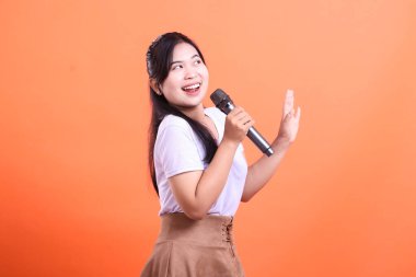 A joyful young woman passionately singing into a microphone, expressing her enthusiasm with an open gesture and radiant smile. isolated on orange background