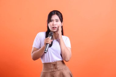A young woman looks stressed and confused while holding a microphone, touching her face, isolated on orange background