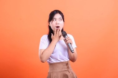 Elinde mikrofon tutan genç bir kadın bir şeye şaşırmış ya da şok olmuş gibi bakıyor. Ağzını turuncu arka planda izole edilmiş, anlamlı bir yüzle kapatıyor.