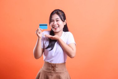 Gülümseyen genç bir Asyalı kadın kredi kartını gösterir ve alışveriş için açık avucuyla sunar. turuncu arkaplanda izole