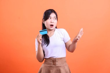 Şaşırmış bir kadın kredi kartını elinde şaşırtıcı bir ifadeyle tutuyor, harcamalarına inanmadığını ve şaşırdığını gösteriyor. turuncu arkaplanda izole
