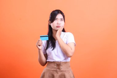 Genç bir kadın, elinde kredi kartı tutarken düşünceli bir ifadeyle yukarı bakıyor, bununla ne yapacağından emin değil gibi görünüyor. turuncu arkaplanda izole