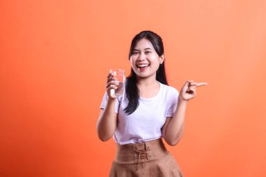 Neşeli Asyalı kadın elinde bir bardak su tutarken mutlu bir ifadeyle yüzünü gösteriyor. Hidrasyon ve sağlıklı yaşam tarzını teşvik etmek, turuncu arka planda izole edilmiş..
