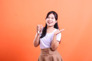 Genç Asyalı kadın bir bardak su sunarak, sağlıklı su ve refahı teşvik ediyor, turuncu arka planda izole edilmiş..
