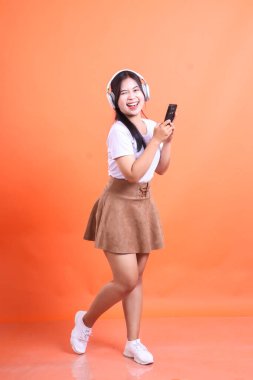 Beyaz tişörtlü, kahverengi etekli, kulaklık takan, akıllı telefon tutan, göz kırpan ve gülümseyen neşeli bir genç kadın. turuncu arkaplanda izole.