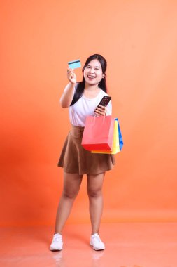 Gülümseyen bir kadın kameraya bakarken bir kredi kartı, alışveriş çantası ve telefon tutar. turuncu arkaplanda izole.