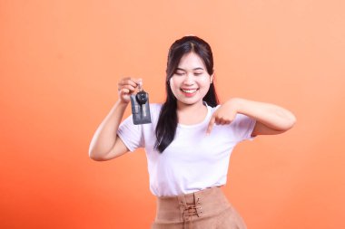 Heyecanlı Asyalı kadın yeni araba anahtarlarını tutuyor ve parmağıyla işaret ediyor, gülümsüyor. Turuncu arka planda izole edilmiş.