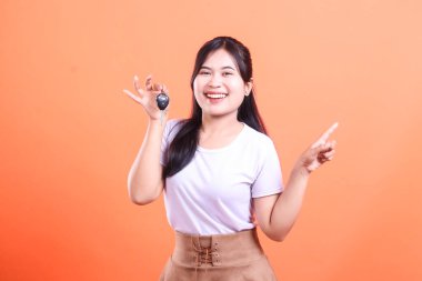 Neşeli bir Asyalı kadın arabanın anahtarını tutuyor ve yan tarafına mutlu bir ifadeyle işaret ediyor. Bu da turuncu arka planda izole edilmiş yeni bir satın alma ya da mülkiyet olduğunu gösteriyor..