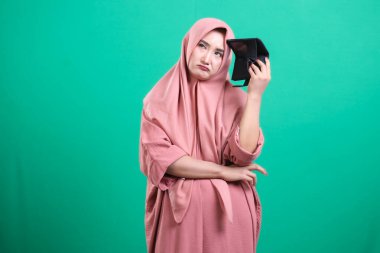 Cüzdanı boş, sıkıntılı bir kadın, parasal zorlukları simgeliyor. Pembe bir başörtüsü takıyor, umutsuzluğunu dile getiriyor, yeşil bir arka planda izole edilmiş. Ekonomik zorluklar, bütçe sorunları, harcamalar ve mali istikrarsızlık.