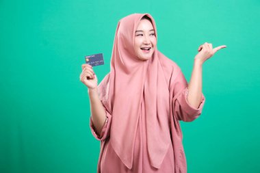 Tesettürlü, tesettürlü, elinde kredi kartı olan ve yeşil arka planda izole edilmiş bir şeyi işaret eden heyecanlı müslüman bir kadın. Bu bir reklam olabilir..