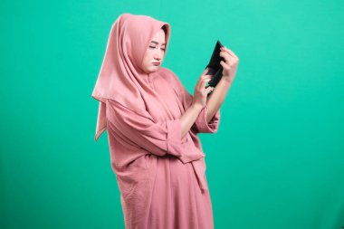 Boş bir cüzdan taşıyan tesettürlü genç müslüman kadın, parasızlıktan rahatsız görünüyor, yeşil bir arka planda izole edilmiş, maddi sıkıntıları ortaya koyuyor..