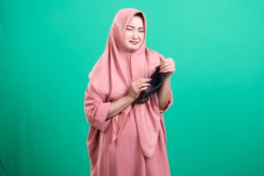 Ümitsiz, tesettürlü Müslüman bir kadın yeşil bir arka planda soyutlanmış ekonomik zorlukların sonuçlarını gösteren boş cüzdanına bakıyor..