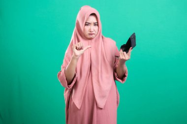 Çaresiz, tesettürlü Müslüman bir kadın, yeşil arka planda izole edilmiş mali zorlukları ve ekonomik zorlukları simgeleyen boş bir cüzdan sergiliyor. Görüntü, yoksulluk ve umutsuzluk hissini taşır..