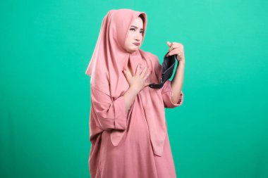 Boş cüzdana bakan tesettürlü üzgün kadın, ekonomik zorlukları ve finansal zorlukları sembolize ediyor, yeşil arka planda izole edilmiş. Para sıkıntısı yüzünden stresli görünüyor..