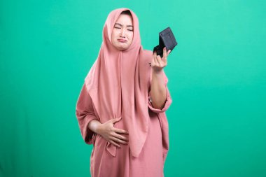 Başörtüsü içindeki üzgün müslüman kadın yeşil arka planda izole edilmiş, maddi sıkıntıları ve ekonomik zorlukları simgeleyen boş bir cüzdan sergiliyor. Yoksulluğun ve çaresizliğin görsel temsili, yeşil bir arka planda izole edilmiş..