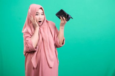 Başörtüsü takan kadın, yeşil arka planda izole edilmiş mali zorlukları temsil eden şok ifadesiyle boş cüzdanına bakar..