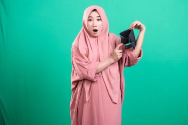 Başörtüsü içindeki perişan bir kadın, yeşil arka planda izole edilmiş, zor zamanlardaki maddi sıkıntıların yaygın sorununu simgeleyen boş bir cüzdan sergiliyor. Küresel ekonomik zorluklarla karşı karşıya.