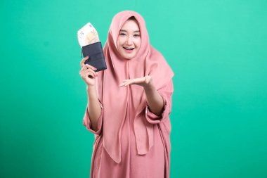 Elinde dolu bir cüzdan olan tesettürlü gülümseyen müslüman kadın yeşil bir arka planda izole edilmiş bir şekilde bir avuç dolusu hediye veriyor. Modern tarzda finansal konsept.