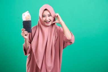 Pembe tesettürlü neşeli Müslüman kadın elinde bir cüzdan dolusu avro para ile kalp hareketi gösteriyor. Yeşil arka planda izole edilmiş finansal refah kavramı.