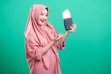 Cüzdanı rupiah dolu, parası olan neşeli Müslüman kadın yeşil bir arka planda izole edilmiş mali bolluk ve refahı temsil ediyor. Başarılı yatırımları ve gelecekteki tasarrufları sembolize eder.