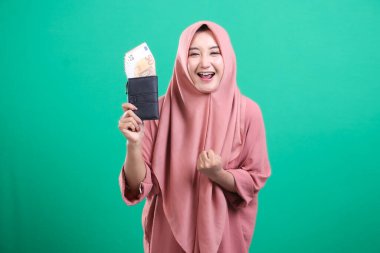 Başörtüsü takan neşeli bir kadın para dolu bir cüzdan taşıyor, yeşil bir arka planda izole edilmiş mutluluk ve finansal güvenlik yayıyor. Finansal zaferini muzaffer bir ifadeyle kutluyor..