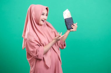 Avro dolu bir cüzdan sergileyen mutlu Müslüman kadın, yeşil bir arka planda izole edilmiş mali özgürlük ve başarıyı simgeliyor. Modern İslami finans sektöründe refah, ekonomik bağımsızlık ve tasarruf göstergesi.