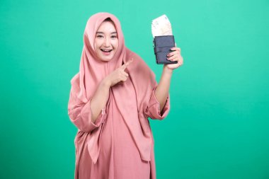 Heyecanlı tesettürlü Müslüman kadın, yeşil arka planda izole edilmiş mali bolluğunu ve başarısını sergileyerek cüzdanını paralarla işaret ediyor. Bu refah ve ekonomik büyümeyi temsil eder.