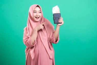 Pembe tesettürlü keyifli genç Müslüman bir kadın cüzdanını parayla dolduruyor ve onu işaret ediyor, yeşil bir arka planda izole edilmiş, neşe dolu..