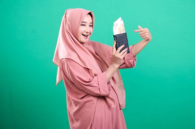 Banknotlarla dolu bir cüzdan taşıyan tesettürlü Müslüman bir kadın, yeşil bir arka planda izole edilmiş, olumlu bir ifadeyle parayı işaret ediyor..