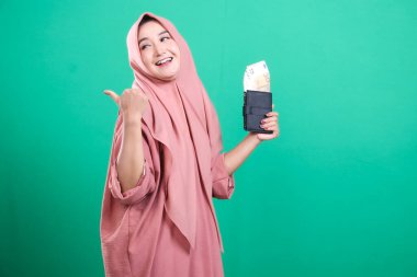 Avro banknotlarıyla cüzdanını tutan Müslüman bir iş kadını, yeşil arka planda izole edilmiş olumlu bir jest yapıyor. Finansal başarı ve ekonomik refahı gösteriyor.