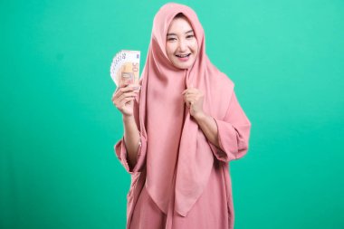 Avro parasının tesettürlü mutlu Müslüman kadın portresi, yeşil bir arka planda izole edilmiş mali refah ve başarı sergiliyor. Yatırım ve kazanç fırsatı sunar.