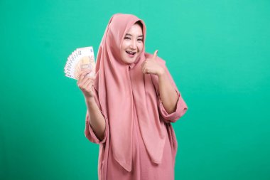 Çekici Endonezyalı kadın tesettür takıyor, sevinçle avro banknotlarını gösteriyor ve yeşil arka planda izole edilmiş, parasal başarıyı işaret eden bir jest yapıyor..