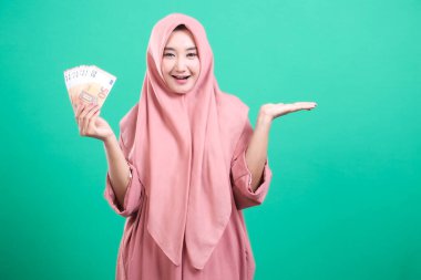 Bir elinde avro banknotları tutan tesettürlü gülümseyen Müslüman kadın, diğer elinde yeşil bir arka planda izole bir şekilde el kol geziyor. Yeşil arka planda izole edilmiş finansal başarı ve tasarrufların desteklenmesi.
