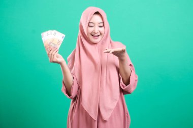 Tesettürlü neşeli Müslüman kadın, yeşil bir arka planda izole edilmiş, yüzünde mutlu bir ifadeyle sunduğu bir jestle avro banknotlarını tutuyor. Bu portre zenginliği ve yatırım başarısını simgeliyor..