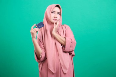 Başörtüsü takan düşünceli Müslüman bir kadın, yeşil bir arka planda izole edilmiş mali kararlar veya işlemler düşünüyor gibi görünen bir kredi kartı tutuyor. İfadesi, çevrimiçi ticaret, alışveriş ve ödemelerle ilgili belirsizliği iletiyor.