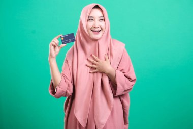 Başörtüsü takan, elinde kredi kartı tutan, yeşil arka planda izole edilmiş modern finansal güç ve neşeyi somutlaştıran keyifli bir kadın. Gülüşü online alışveriş ve güvenli bankacılık işlemlerinin kolaylığını iletiyor.