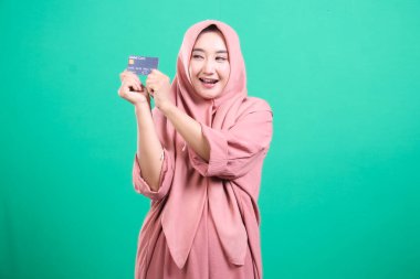 Hevesli Müslüman kadın, yeşil arka planda izole edilmiş modern finans sembolize ederek banka kartı sergiliyor. Neşeli ifadesi finansal bir güçlenme hissi veriyor..