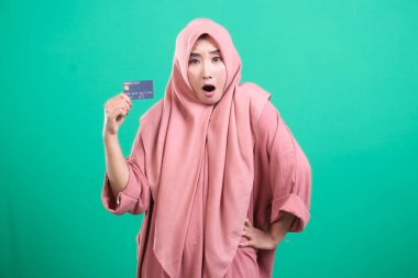 Çekici Müslüman bir kadın, elinde kredi kartı tutarken şok olmuş görünüyor. Sanki yeşil arka planda beklenmedik harcamalar yapıyormuş gibi..