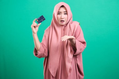 Tesettürlü tesettürlü Müslüman bir kadın kredi kartı tutuyor ve yeşil arka planda izole edilmiş endişe ve mali kaygılarını dile getiriyor. Resim borcun yükünü betimliyor..