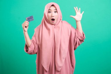 Tesettürlü genç Müslüman bir kadın kredi kartı gösteriyor. Şaşırmış görünüyor. Finansal konsept, yeşil arka planda izole edilmiş alışveriş..