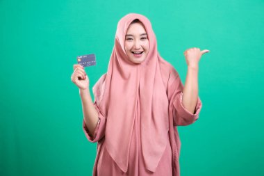 Neşeli Müslüman kadın tesettür takıyor ve banka kartı tutuyor, yeşil arka planda izole edilmiş finansal işlemlerde kolaylık ve ulaşılabilirlik sunuyor, modern İslami yaşam tarzını memnuniyetle yansıtıyor.