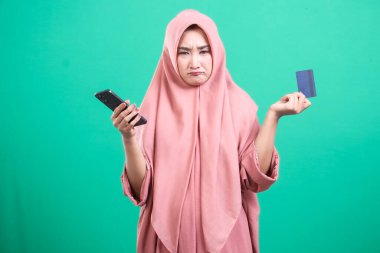 Başörtüsü takan genç Müslüman bir kadın, yeşil bir arka planda izole edilmiş, telefon ve kredi kartını kullanarak online ödeme sorunlarıyla boğuşurken, kafası karışık ve üzgün görünüyor..
